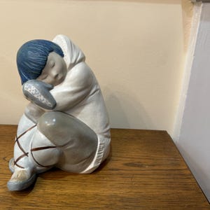Peut inclure: Une figurine en porcelaine d'une personne assise la tête baissée, portant une veste à capuche blanche, un pantalon bleu à rayures marrons et un chapeau bleu. La figurine est en position assise, les bras enroulés autour des jambes.