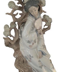 May include: A porcelain figurine of a woman in a white kimono, holding a fan, seated near a tree branch. The figurine has grey hair and is decorated with floral patterns in soft pastel colours. The tree branch is a light brown colour.