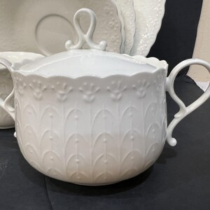 May include: White porcelain sugar bowl with a lid and two handles. The bowl features a decorative embossed pattern of stylized leaves and a scalloped rim. Matching saucer and other dishware are visible in the background.