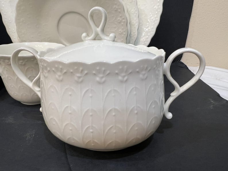 May include: White porcelain sugar bowl with a lid and two handles. The bowl features a decorative embossed pattern of stylized leaves and a scalloped rim. Matching saucer and other dishware are visible in the background.