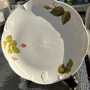 Può includere: Una ciotola da portata in ceramica bianca con un bordo ondulato e un motivo floreale. La ciotola presenta una farfalla, boccioli di rosa e foglie in tonalità di verde, marrone e rosso.