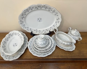 May include: A white porcelain dinner service with a grey floral design. The set includes an oval serving platter, serving bowls, plates, a teacup and saucer, a gravy boat, and a sugar bowl. The edges are scalloped and silver-trimmed.