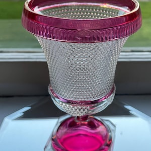 May include: A clear glass vase with a pink rim and base. The vase has a textured, dotted surface and a flared, trumpet-shaped body.