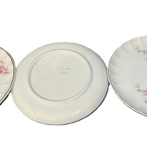 May include: Three white ceramic plates with a floral design. Two plates are oval-shaped with a gold rim and a pink cherry blossom pattern. The third plate is round, with the underside visible.