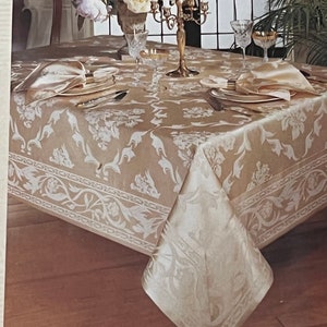 Peut inclure: Une nappe beige avec un motif floral blanc et une bordure de vignes blanches. La nappe est dressée avec un centre de table composé d'un chandelier doré et de fleurs blanches dans un vase. Il y a deux couverts avec des serviettes blanches et des couverts en argent.