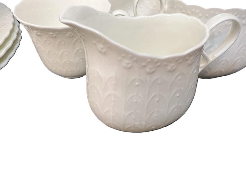 May include: A set of white porcelain serving items, including a creamer, bowls, and plates. The creamer features a handle and an embossed decorative pattern. The items are arranged on a white background, suitable for a formal dining occasion.