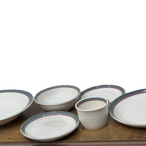 May include: A collection of white ceramic dishware with a teal and maroon border. The set includes bowls, plates, and a cup, arranged on a wooden surface. Ideal for serving meals.