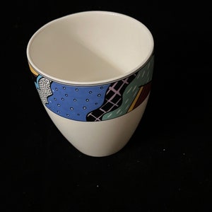 May include: White ceramic mug with a colorful abstract design featuring blue, green, yellow, and black. The design is a geometric pattern with a black outline.