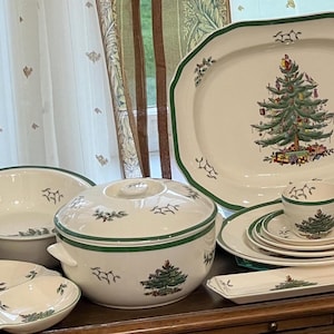 Op de afbeelding: Een verzameling groene en witte kerstboom-gepatroneerde keramische schalen, waaronder een theepot, een serveerschaal, een ovenschaal en verschillende borden. De schalen zijn gerangschikt op een houten tafel.
