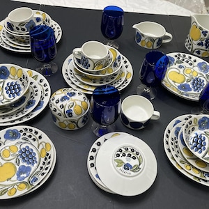 May include: A collection of white ceramic tableware with a floral and fruit design in blue and yellow. Includes plates, bowls, cups, saucers, a creamer, and a pitcher. Also, cobalt blue stemmed glasses.