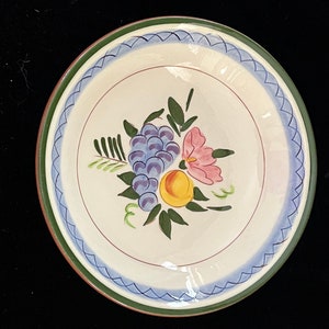 May include: A white ceramic bowl with a green, blue, and red rim. The bowl features a hand-painted design of a pink flower, a yellow peach, and a bunch of purple grapes.