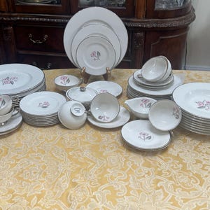 Op de afbeelding: Een set wit porselein serviesgoed met een delicaat bloemenmotief in roze en grijs. De set bevat borden, kommen, kopjes en schoteltjes.