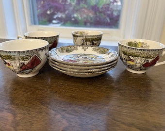Può includere: Un set di quattro tazze da tè e piattini vintage con un design a tema invernale. Le tazze e i piattini sono bianchi con un motivo marrone, verde e rosso. La scena rappresenta un paesaggio innevato con case e alberi.
