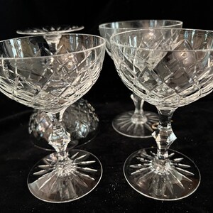 May include: Four clear crystal champagne coupes with a diamond pattern. The coupes have a wide bowl and a long stem with a starburst design at the base.