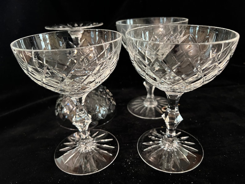 May include: Four clear crystal champagne coupes with a diamond pattern. The coupes have a wide bowl and a long stem with a starburst design at the base.