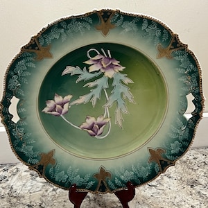 May include: A vintage green and gold porcelain plate with a floral design. The plate has two handles and a scalloped edge.