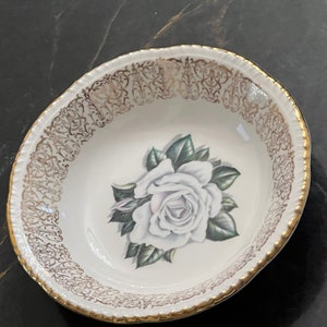 May include: Oval-shaped white ceramic bowl with a gold rim and decorative gold filigree border. The bowl features a central image of a white rose with green leaves. A vintage-style decorative piece.