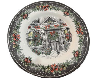 May include: A white ceramic plate with a black dotted border and a detailed Christmas scene. The plate features a house with a wreath on the door, snow, and red bows. The border has bells, pinecones, and bows.