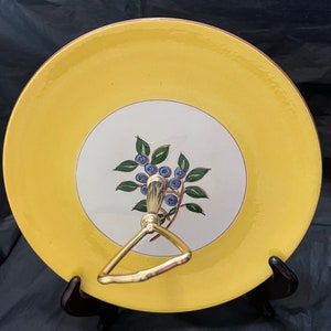 May include: A yellow ceramic serving plate with a gold rim and a white center featuring a painted design of blueberries on a branch. The plate has a gold metal handle.