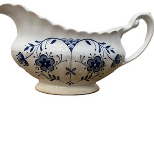 以下が含まれることがあります： 白のセラミック製グレービーボート。縁はスカラップで、取っ手が付いています。青い花柄とつる柄で装飾されています。注ぎ口があり、小さな丸い台座の上に置かれています。クラシックな食器です。