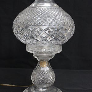 May include: A clear glass table lamp featuring an intricate diamond-cut pattern across its bulbous body and circular base. The lamp has a flared top opening and a central stem connecting the base to the main body, with a visible electrical cord.