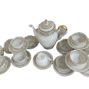 May include: A white porcelain tea set with gold trim. The set includes a teapot, creamer, sugar bowl, and multiple teacups and saucers. The teapot has a decorative lid and gold accents on the handle and spout. The teacups and saucers are arranged in a neat display.
