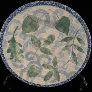 Puede incluir: Un plato de cerámica blanco con un patrón de hojas azules y verdes. El plato tiene un borde azul decorativo con un diseño de espiral.