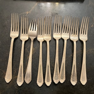 Può includere: Un set di dieci forchette d'argento con disegni decorativi. Le forchette sono disposte in fila, con i denti rivolti verso l'alto.