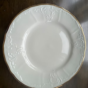 May include: A white porcelain plate with a gold rim and ornate embossed floral and scrollwork detailing. The plate has a scalloped edge and a slightly recessed center, suitable for serving food.