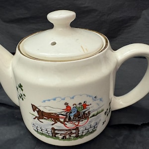 Puede incluir: Tetera de cerámica blanca con ribete dorado y una escena de un carruaje tirado por un caballo con personas dentro. La escena está pintada en tonos de azul, verde, rojo y marrón.