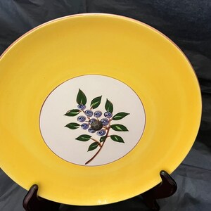 May include: A yellow ceramic plate with a white center featuring a hand-painted design of a blueberry branch with green leaves and blue berries.