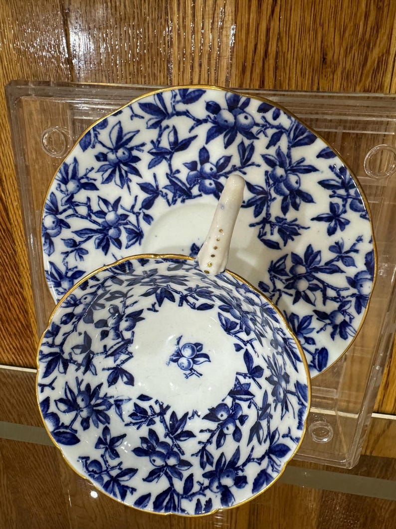 Może przedstawiać: Zestaw filiżanki i spodeczka do herbaty z porcelany w kolorze niebieskim i białym z pozłacanym rantem. Zestaw zdobi kwiatowy wz&oacute;r z niebieskimi jagodami i liśćmi.