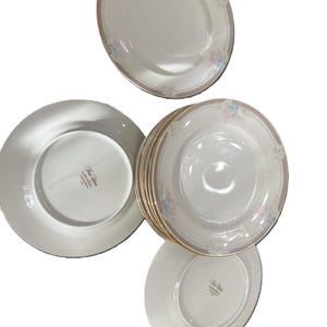 May include: A set of white ceramic plates featuring a delicate floral pattern in pastel pink and blue along the rim, accented with a thin gold band. The underside of the plates displays the text Fine China J-MICASA L-9000 JAPAN.