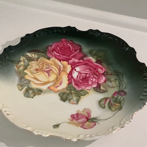 May include: A white porcelain plate with a green and white floral design. The plate features a large yellow rose with pink roses and green leaves. The plate has a scalloped edge.