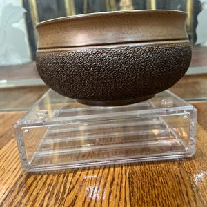 May include: A brown ceramic bowl with a textured surface sits on a clear acrylic stand. The bowl has a slightly rounded shape and a wide rim.