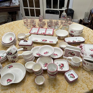 May include: A large collection of cream-colored ceramic dinnerware featuring a red apple pattern with green leaves. The set includes various bowls, plates, serving dishes, a butter dish, a creamer, a napkin holder, and several drinking glasses.