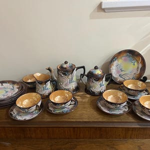 Op de afbeelding: Een vintage theeservies met een drakenontwerp. De set bestaat uit een theepot, een roomkan, een suikerpot en zes theekopjes en schoteltjes. De theekopjes en schoteltjes zijn versierd met een witte draak op een bruine achtergrond.