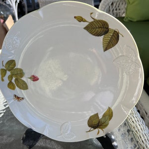 May include: White ceramic dinner plate with a scalloped edge and a floral design featuring green leaves, pink roses, and a butterfly.