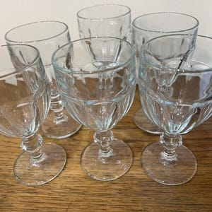 May include: Six clear wine glasses with a textured, ridged pattern on the bowl. The glasses have a classic shape with a round base and a detailed stem. They are arranged on a wooden surface.