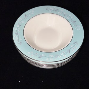 May include: A stack of white ceramic bowls with a pale blue rim. The rim features a floral pattern of small white flowers and grey leaves. The bowls have a silver trim.