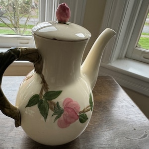 May include: Cream-colored ceramic teapot with a rose motif. The teapot features a curved spout, a handle designed to resemble a branch, and a lid topped with a pink rosebud. Pink roses and green leaves are painted on the body.