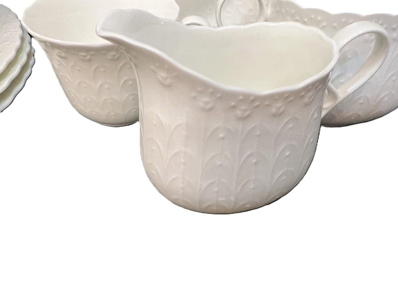 May include: A collection of white porcelain serving dishes. The set includes a creamer with a handle, a bowl, and a plate. Each piece features a raised, repeating leaf and dot pattern.