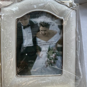 May include: A white photo frame with a decorative floral design and a gold trim. The frame holds a black and white photo of a wedding couple. The text "OPAL INNOCENCE" and "LENOX" are visible on the frame.
