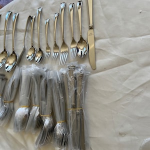 Puede incluir: Una colección de cubiertos de acero inoxidable, que incluye cucharas, tenedores y cuchillos, dispuestos sobre una superficie blanca. Algunas piezas están sueltas, mientras que otras están agrupadas y envueltas en plástico. Los cubiertos tienen un acabado pulido y reflectante.