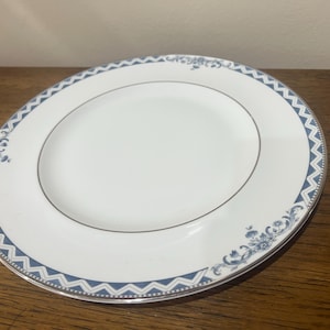 May include: A white ceramic dinner plate with a blue and silver patterned border. The border features a chevron design and floral accents.