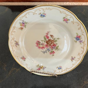 May include: A cream-coloured porcelain plate with a gold rim and floral design. The plate features a central bouquet of pink, brown, and blue flowers, with smaller floral clusters around the scalloped edge. The plate is approximately 25 cm in diameter.