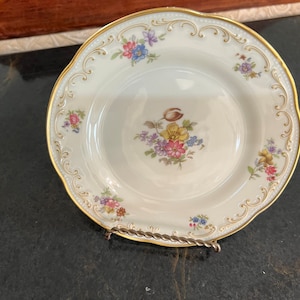 Puede incluir: Un plato de porcelana blanca con un borde dorado y un diseño floral. El plato presenta un motivo floral central con flores rosas, amarillas y azules. El plato está sobre un soporte decorativo.