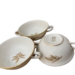 May include: Three white porcelain soup bowls with gold trim and wheat stalk designs. Each bowl has two handles and a gold rim. The bowls are arranged in a slightly overlapping pattern.