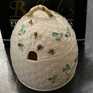 May include: White ceramic honey pot shaped like a beehive with a lid and a small opening for pouring. The pot is decorated with green shamrocks and brown bees.