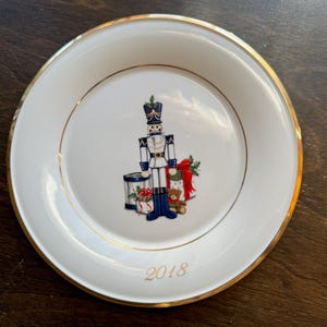 Könnte beinhalten: Ein weißer Keramikteller mit goldfarbenem Rand, der eine weihnachtliche Illustration eines Spielzeugsoldaten, einer Trommel und eines Teddybären zeigt. Der Teller ist mit einem goldenen Rand verziert und das Jahr "2018" ist in Gold auf der Unterseite aufgedruckt.